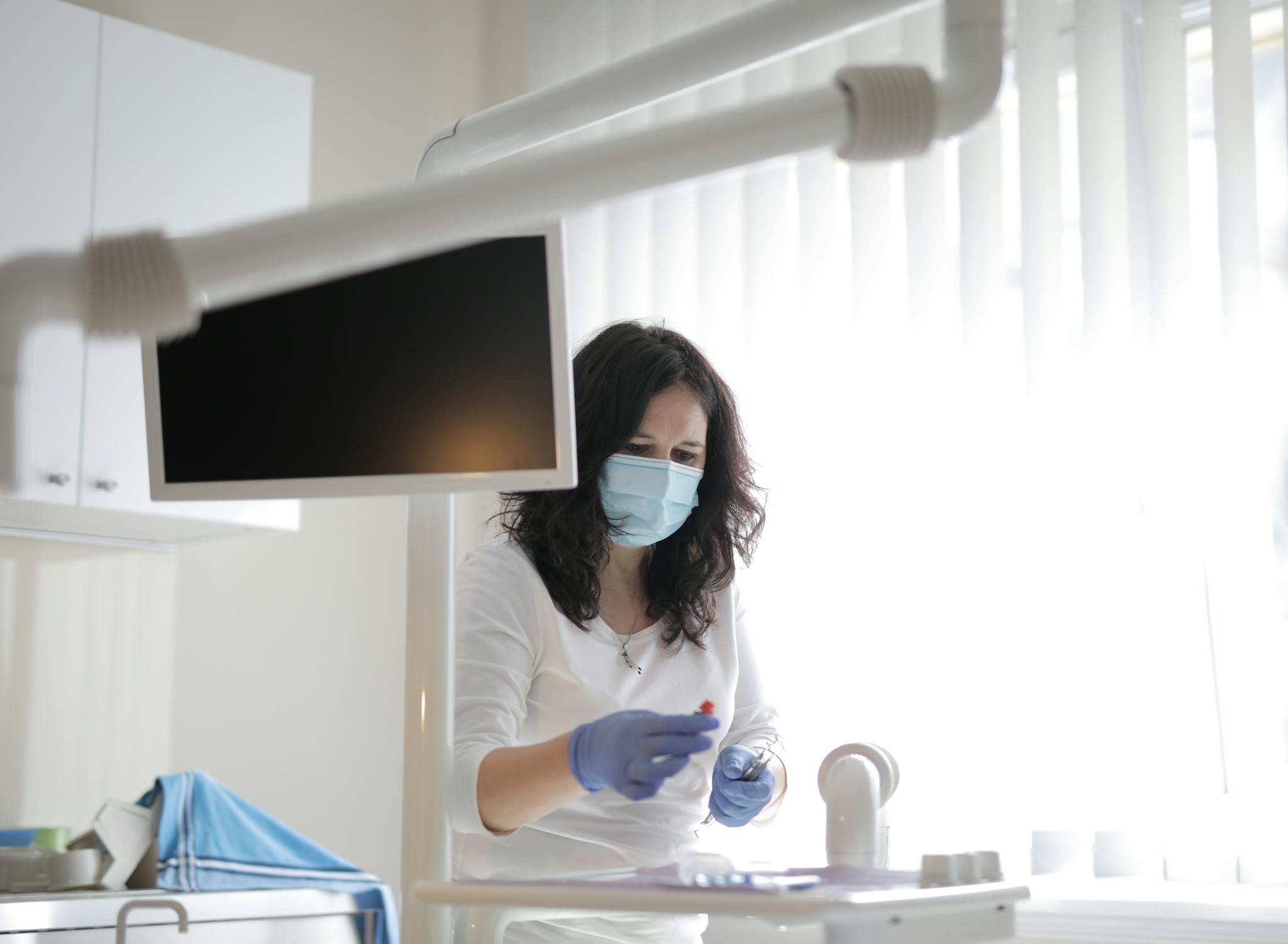 Serious female dentist in medical mask and latex gloves wearing white uniform preparing instruments for treatment in light modern dental room in clinic